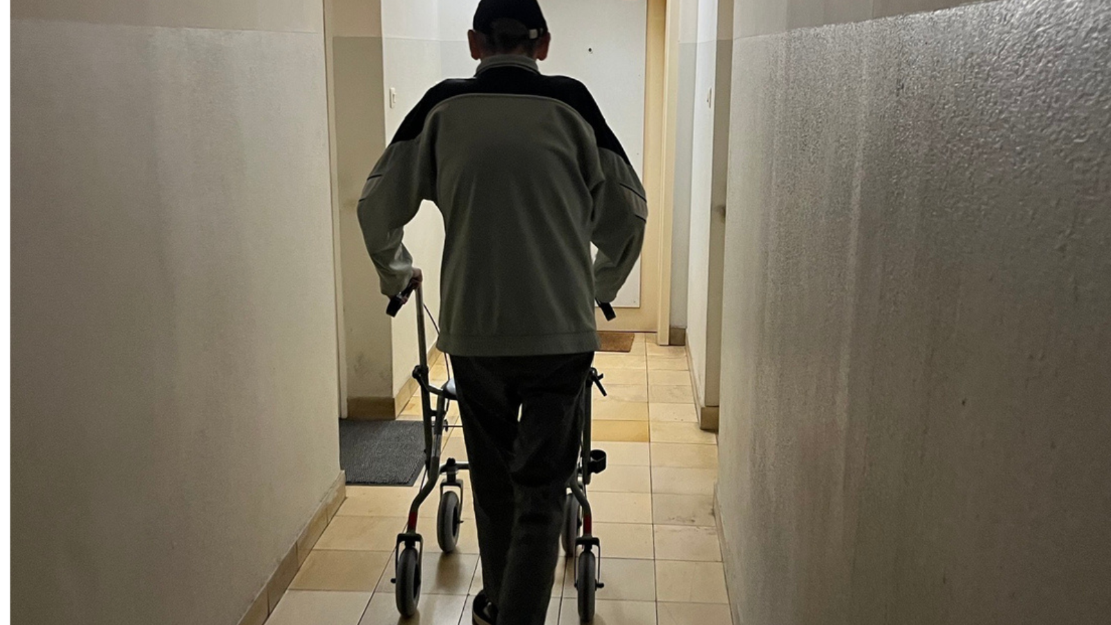 Ein älterer Mann mit einem Rollator ist von hinten in einem schwach belichteten Flur zu sehen.